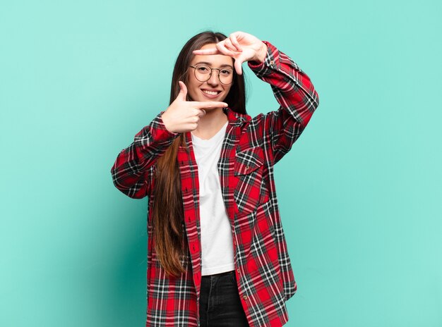 Young pretty casual woman feeling happy, friendly and positive, smiling and making a portrait or photo frame with hands