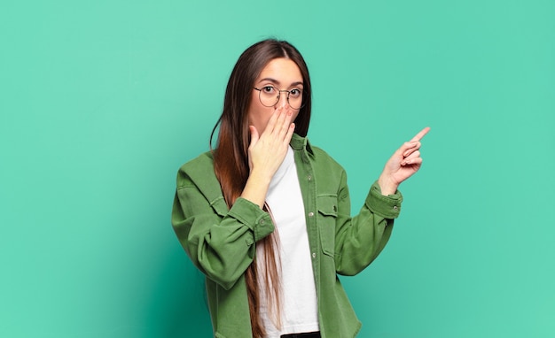 Young pretty casual woman feeling happy, shocked and surprised, covering mouth with hand and pointing to lateral copy space