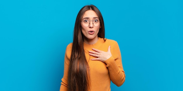 Young pretty casual woman feeling shocked and surprised, smiling, taking hand to heart, happy to be the one or showing gratitude