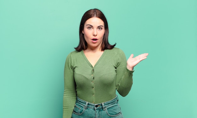 Young pretty casual woman looking surprised and shocked, with jaw dropped holding an object with an open hand on the side