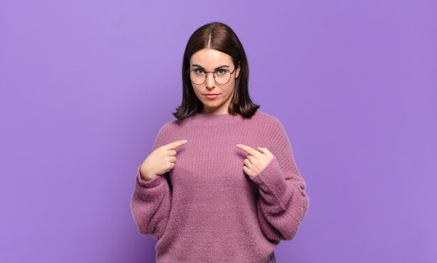 Young pretty casual woman pointing to self with a confused and quizzical look, shocked and surprised to be chosen