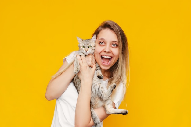 A young pretty caucasian excited smiling blonde woman in a white tshirt with a cat in her hands is happy about the news isolated on a bright color yellow wall