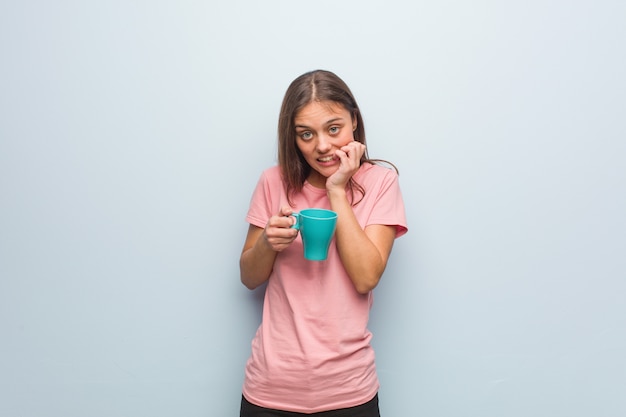 Young pretty caucasian woman biting nails, nervous and very anxious. She is holding a mug.