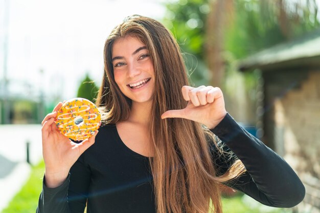 Young pretty caucasian woman holding a donut at outdoors proud and selfsatisfied