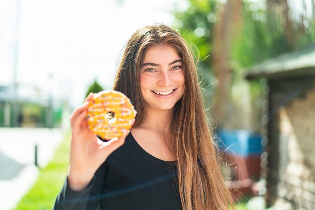 Young pretty caucasian woman holding a donut at outdoors with happy expression