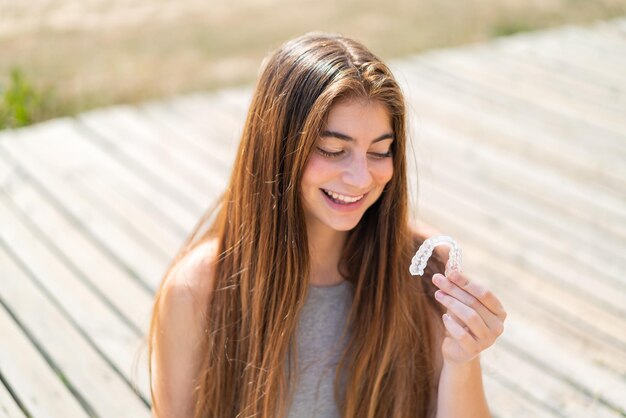 Young pretty caucasian woman holding invisible braces at outdoors with happy expression