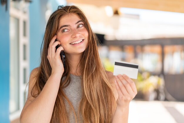 Фото Молодая красивая белая женщина ведет разговор с мобильным телефоном и держит кредитную карту