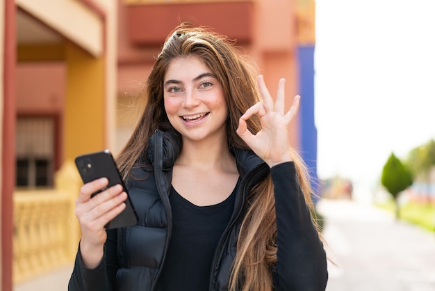 Молодая красивая белая женщина использует мобильный телефон на открытом воздухе, показывая знак "ок" пальцами