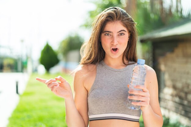 Молодая красивая белая женщина с бутылкой воды на открытом воздухе удивлена и указывает на сторону