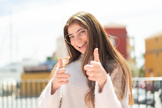 Foto giovane bella donna caucasica con gli occhiali all'aperto che indica la parte anteriore e sorride