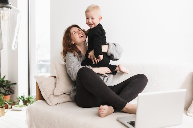 写真 彼女の小さな笑う息子と一緒に楽しく遊んでソファに座っている若いかなり陽気な女性