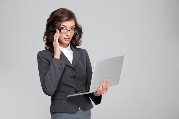 Young pretty concentrated confident businesswoman holding laptop and touching her glasses