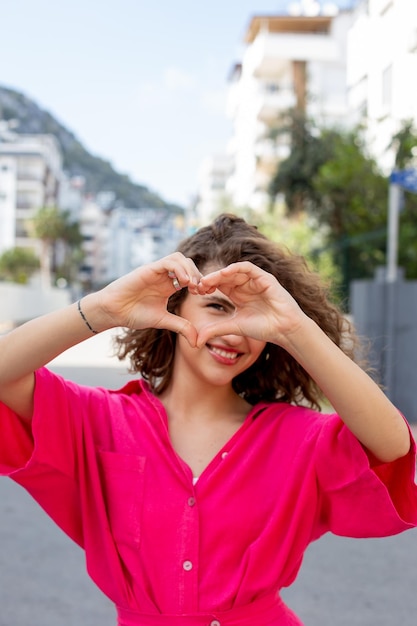 Foto giovane donna dai capelli ricci che fa la forma di un cuore con le mani per le strade della città ama le emozioni