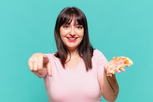 Молодая симпатичная пышная женщина, указывающая или показывающая и держащая пиццу