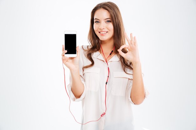 Молодая довольно милая девушка с наушниками, слушающая музыку со смартфоном и показывающая жест, изолированный на белой стене