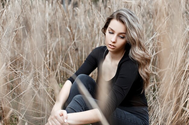 Giovane donna abbastanza carina in jeans alla moda in maglietta nera alla moda sta riposando seduto per terra nel campo