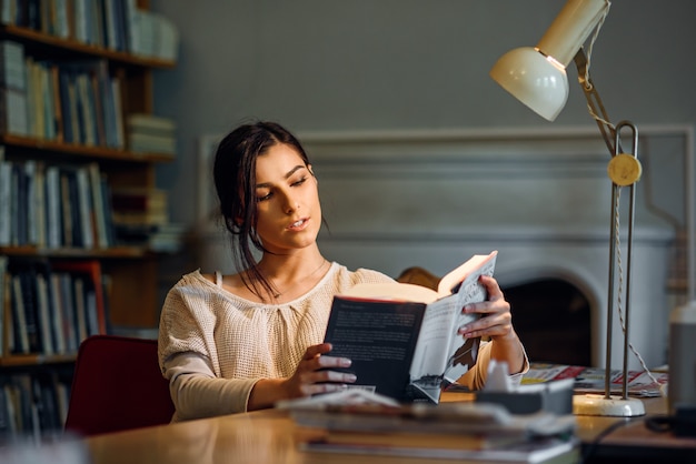 Giovane libro di lettura grazioso ed entusiasta della ragazza dello studente in biblioteca universitaria sotto una lampada da tavolo