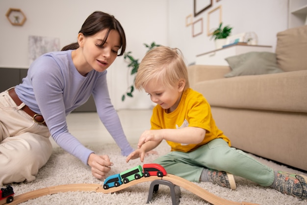 Young pretty female and adorable little blond boy sitting on fluffy rug on the floor by couch and playing with toy train together at home