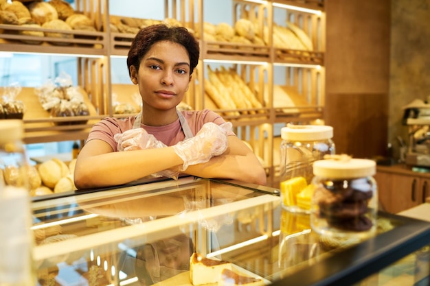 Young pretty female clerk standing by glass display with pastry