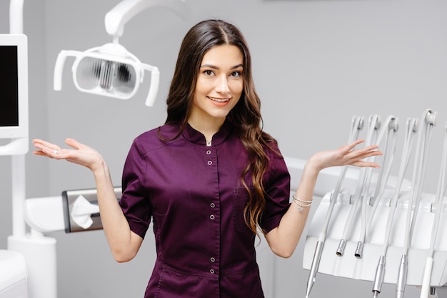 A young pretty female dentist is standing near the dental chair in the office gesturing with her hands