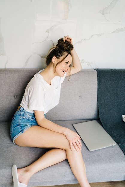 молодая симпатичная женщина делает покупки в Интернете на ноутбуке, она лежит на диване и печатает на ноутбуке