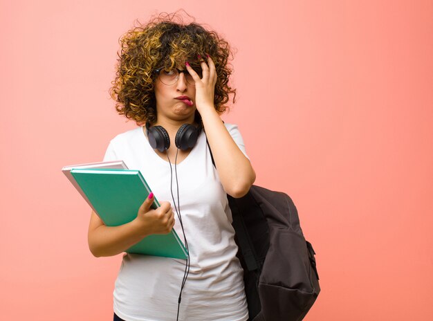 Young pretty female student feeling bored, frustrated and sleepy after a tiresome, dull and tedious task, holding face with hand over pink wall