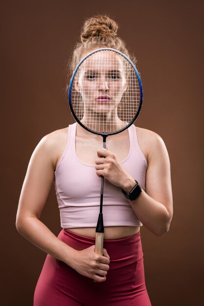 Молодая симпатичная теннисистка в розовой майке и малиновых леггинсах держит ракетку перед лицом
