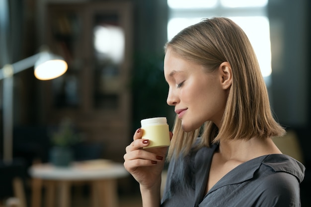 Young pretty female with blond hair enjoying smell of natural handmade cosmetic product in small plastic jar before applying it on her skin