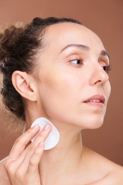 Young pretty female with cotton pad cleaning her neck with purifying lotion while taking care of face and body in the morning