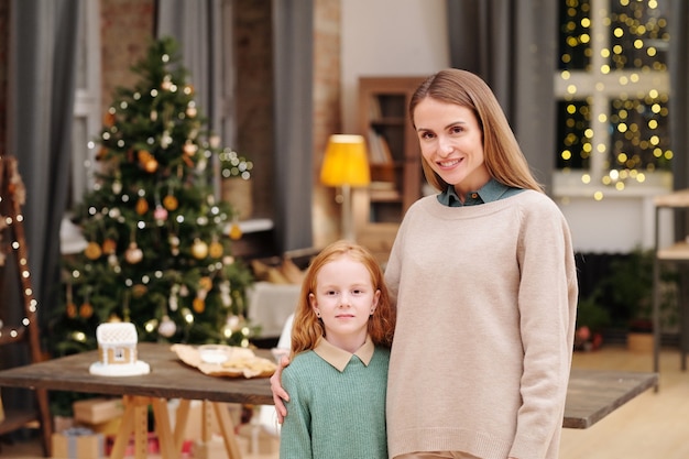 Young pretty female with toothy smile embracing her cute little daughter while both standing in front of camera against decorated firtree at home