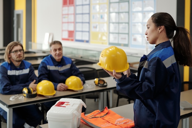 Giovane bella lavoratrice in uniforme in piedi di fronte ai suoi colleghi maschi mentre descrive lo scopo dell'uso del casco protettivo