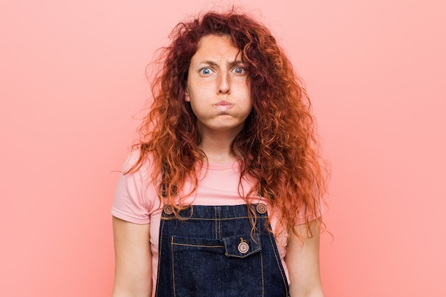 La giovane donna graziosa della rossa dello zenzero che indossa un salopette dei jeans soffia le guancie, ha espressione stanca. concetto di espressione facciale