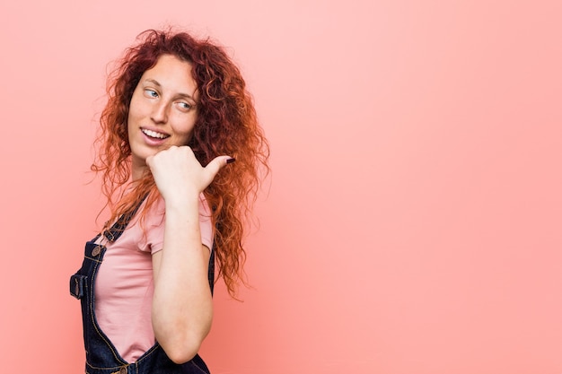 Young pretty ginger redhead woman wearing a jeans dungaree points with thumb finger away, laughing and carefree.