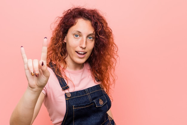 Young pretty ginger redhead woman wearing a jeans dungaree showing a horns gesture