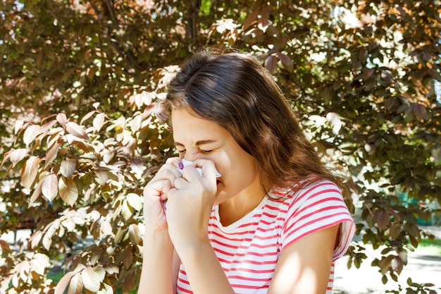 Young pretty girl blowing nose in front of tree. Spring allergy concept