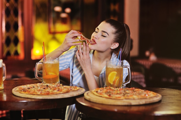 A young pretty girl eating pizza and drinking beer or a beer citrus cocktail on the surface of a bar or pizzeria.