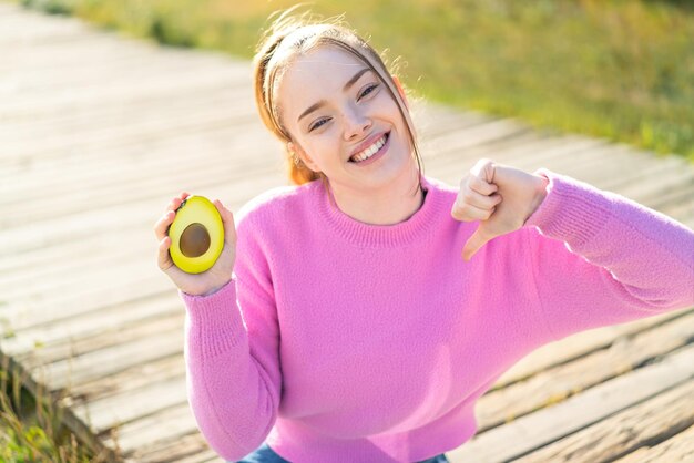Фото Молодая красивая девушка держит авокадо на улице, гордая и самодовольная