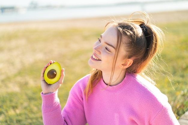 Молодая красивая девушка держит авокадо на улице, улыбаясь и глядя вверх