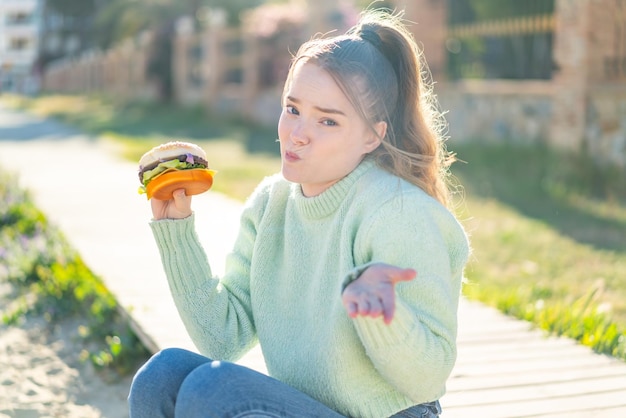 Young pretty girl holding a burger at outdoors making doubts gesture while lifting the shoulders