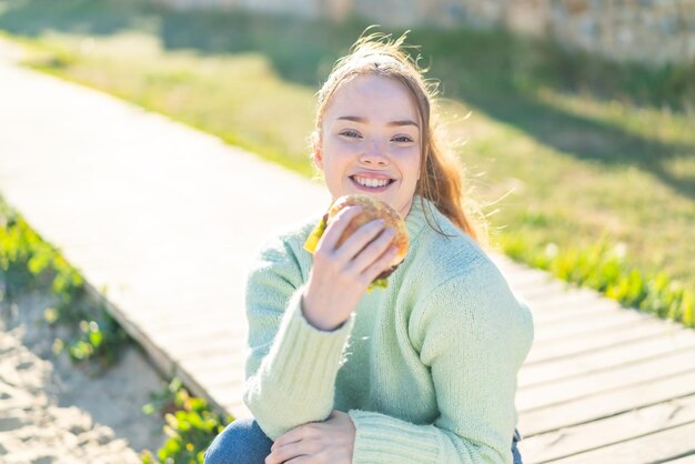 Young pretty girl holding a burger at outdoors with happy expression