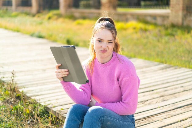 Young pretty girl holding a tablet at outdoors with sad expression