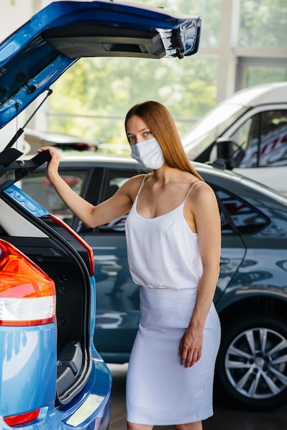 A young pretty girl inspects a new car at a car dealership in a mask during the pandemic. The sale and purchase of cars, in the period of pandemia.