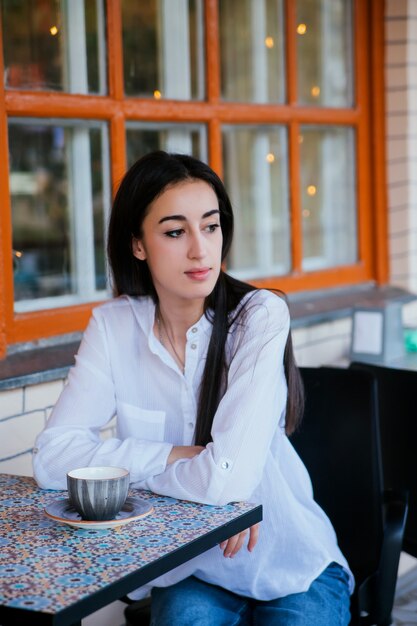 A young pretty girl is sitting at a table in a cafe and drinking coffee or tea.