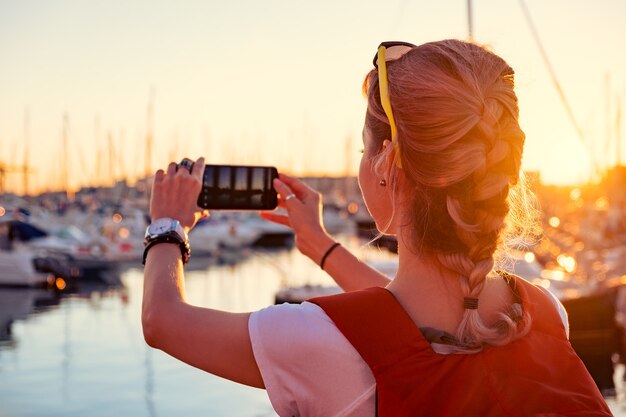 Young pretty girl is taking a picture of a bay with yachts at sunset