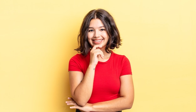 Young pretty girl looking happy and smiling with hand on chin, wondering or asking a question, comparing options