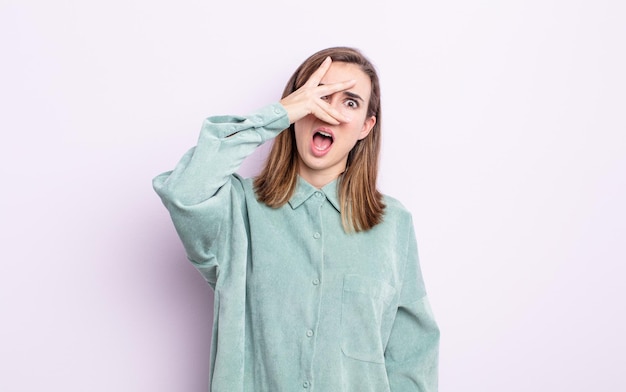 Young pretty girl looking shocked, scared or terrified, covering face with hand and peeking between fingers