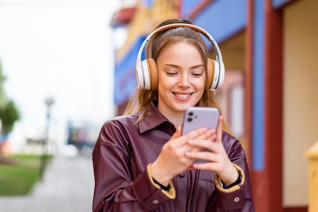 Молодая красивая девушка на улице слушает музыку и смотрит на мобильный