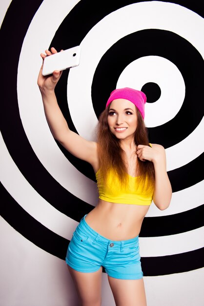 Young pretty girl in pink hat with slim belly making photo on smartphone