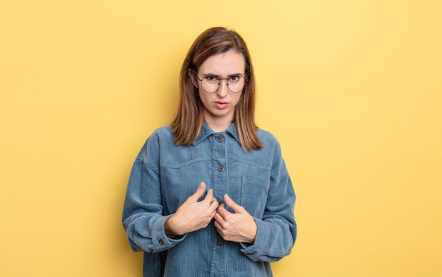 Young pretty girl pointing to self with a confused and quizzical look, shocked and surprised to be chosen