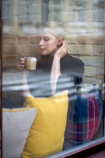 Young pretty girl siting near window and drinking coffee or tea in the cafe. Coffee break.